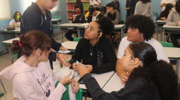 aluna em pé distribui papeis para meninas sentadas em sala de aula. #paratodosverem