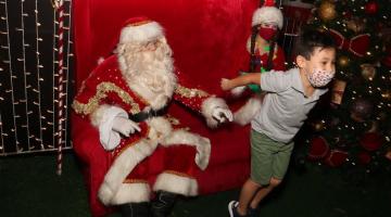 menino sai da cadeira onde está papai noel pulando de alegria. A mamãe noel e uma árvore de natal estão ao fundo. #paratodosverem