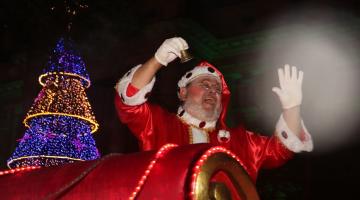 papai noel passa de treno diante de grande árvore de natal iluminada. Ele toca o sino e saúda o público (que não aparece na imagem). Ao fundo, o paço municipal. #paratodosverem