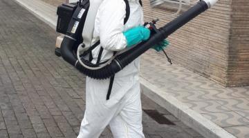 homem está usando equipamento de nebulização do tipo costal, com uma espécie de mochila nas costas ligada a um tubo similar ao de um aspirador de pó. Ele está vestido com macacão, mascara própria para respirar e usa proteção no ouvido. #paratodosverem