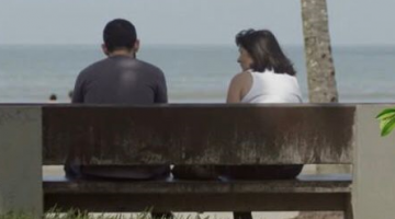 casal está sentado em banco da praia, de frente para o mar e de costas para a foto. #paratodosverem 