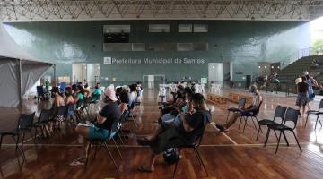 ginásio coberto com cadeira distanciadas e pessoas sentadas. Ao fundo se lê prefeitura municipal de santos. #paratodosverem