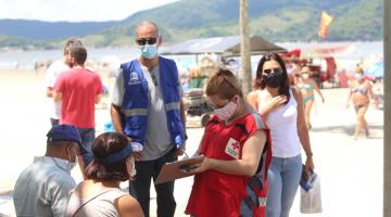 Voluntária da Cruz Vermelha, em pé, conversa com casal sentado em banco e anota dados em prancheta. #pracegover