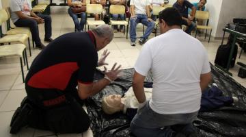 dois homens ajoelhados no chão simulam atendimento a  pessoa ferida. Trata-se de um boneco deitado também no chão. Ao fundo, os alunos atentos. #paratodosverem