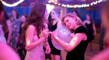 Cena de O Mau Exemplo de Cameron Post. Duas meninas estão num salão onde se passa uma festa. Uma delas está abraçada a um balão de gás. O ambiente está decorado com luzes. Há outra meninas ao fundo. #Pracegover