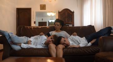 Dois jovens estão deitados em um sofá, de lados opostos, ambos com a cabeça no colo de uma mulher. #Paratodosverem
