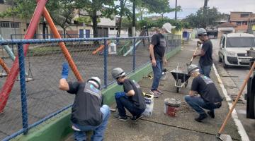 homens trabalham em obra em calçada ao lado de play ground com alambrado. São cinco pessoas, três estão agachadas junto a latas de tinta, pá e carrinho de mãos. Eles usam capacete. #paratodosverem