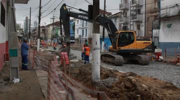 rua com obras. Retroescavadeira em movimento e homens trabalhando. há terra nos canteiros e a pista está coberta  por pedras. #paratodosverem