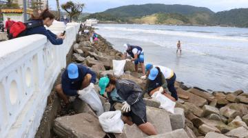 voluntários estão junto às pedras da mureta da ponta da praia, como mar ao fundo. Eles recolhem microlixo. #paratodosverem