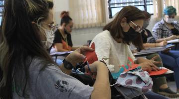 educadoras recortam figuras e desenham em papel. Elas estão sentadas em uma sala de aula. #paratodosverem