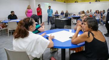 em primeiro plano, mesa com professoras sentadas. Ao fundo, três pessoas em pé, sendo uma delas falando ao microfone. Há outros professores sentados em torno. #paratodosverem 