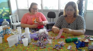 Mulheres estão sentadas e recortando e costurando. Sobre a mesa à frente delas há produtos para artesanato, como tecidos, fitas, linha e tesoura. #Pracegover