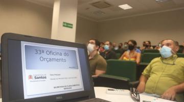 tela de notebook aberta sobre uma mesa em primeiro plano. Na tela se lê 33ª Oficina do Orçamento. O logo da cidade está abaixo. Ao fundo, pessoas sentadas no auditório