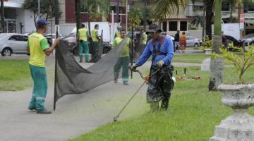 homens estão cortando grama no jardim da praia. Em primeiro plano, um deles usa uma máquina para cortar a grama. Ao fundo, dois seguram uma rede de proteção para a grama cortada não espalhar. #paratodosverem