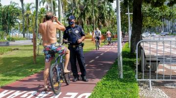 Guarda Municipal orienta ciclista na ciclovia a colocar máscara. O ciclista está de costas para a imagem. Ele leva as mãos à altura da cabeça para colocar o acessório.#Paratodosverem