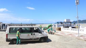 Homens trabalham próximo ao canal, fazendo manutenção. Há um carrinho de mão e um veículo de apoio na areia.#Paratodosverem