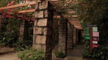 entrada do orquidário com pergolado coberto por vegetação e flores. Ao lado direito está afixada uma placa de orientação. #paratodosverem