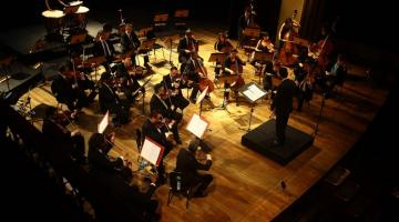 Músicos da orquestra tocam em palco com maestro à frente. #paratodosverem