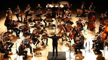 Músicos da orquestra são regidos pelo maestro Luiz Petri, de costas e ao centro da imagem. #pratodosverem. 