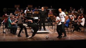 músicos da orquestra tocando e quatro pessoas encenando a disputa de um jogo de futebol no palco. #paratodosverem