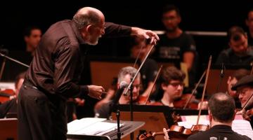 maestro em primeiro plano está regendo orquestra ao fundo. #Paratodosverem