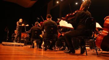 músicos sentados tocando com maestro ao fundo num teatro. #paratodosverem 