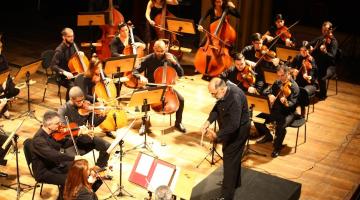 maestro está à frente da orquestra. Todos estão no palco. #paratodosverem