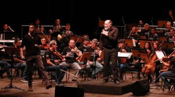 maestro está à frente da orquestra. Ele olha para a frente. Músicos estão atrás. #paratodosverem