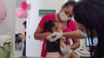 Criança recebe vacina com mãe segurando #paratodosverem