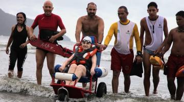 Mulher está sentada em cadeira anfíbia, na beira d'água. Ela está ladeada por um grupo de seis pessoas