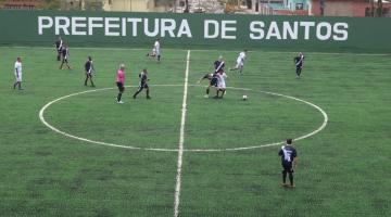 jogo de veteranos no campo renovado. Ao centro, há uma disputa de bola. Ao fundo, se lê: Prefeitura de Santos. #paratodosverem