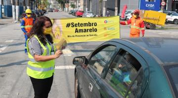 Mulher entrega panfleto a motorista e ao fundo faixa do setembro amarelo #paratodosverem