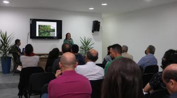 pessoas estão sentadas em auditório de costas para foto. Ao fundo, uma mulher fala ao público. Atrás dela há um monitor com imagem projetada. #paratodosverem