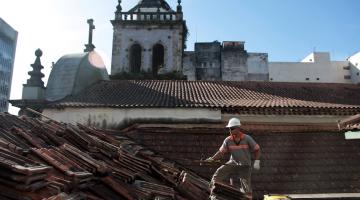 operário está em sobre o telhado de um prédio retirando as telhas. Ao fundo se vê as torres de um conjunto de igrejas. #paratodosverem