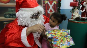 Criança olha curiosa para o papai noel, sentada no colo dele e segurando um presente. #paratodosverem