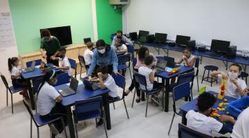 sala com diversas mesas. Alunos sentados mexendo nos computadores. Alguns professores estão ao lado dos alunos. À direita, uma menina monta uma estrutura com peças colorida, como um jogos de peças de encaixe. #paratodosverem