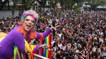 Drag Queen está no alto de um carro alegórico saudando uma multidão em praça. #Pracegover