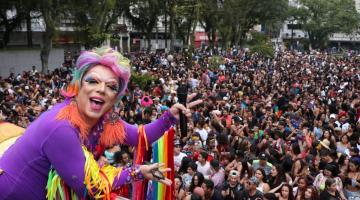 Drag com roupa colorida no alto de carro de som mostra a praça lotada ao fundo. #paratodosverem