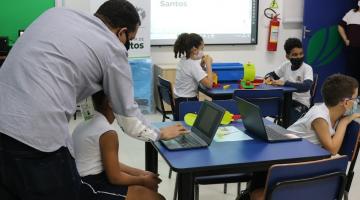 crianças estão sentadas em sala, diante de notebooks. Em primeiro plano, à esquerda, um homem, de costas, tecla no notebook ao lado de uma criança. #paratodosverem