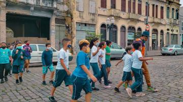 Crianças caminham por rua de paralelepípedo e casas antigas ao fundo. #paratodosverem