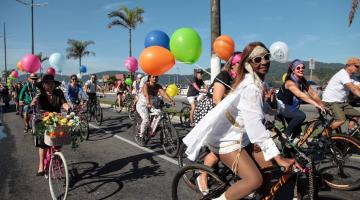 pessoas vestidas com roupas antigas e segurando balões de gás estão montadas em suas bikes e pedalando na avenida da orla. #paratodosverem