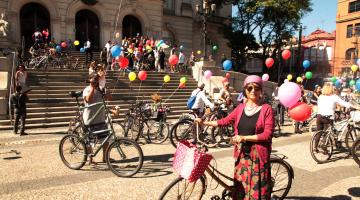 Ciclistas ocupam praça e a escadaria da Praça Mauá. A maioria das bicicletas está decorada com balões coloridos. Em primeiro plano há uma mulher vestida à moda antiga. Ela está em pé à frente de sua bike. #Pracegover