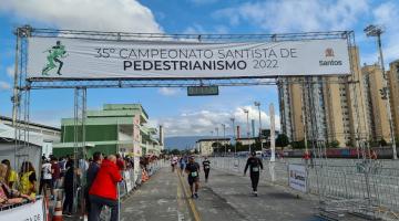 atletas estão chegando ao final da prova. Há um pórtico no alto onde se lê 35º Campeonato Santista de Pedestrianismo. #paratodosverem