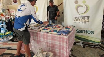 mesa com pacotes de alimentos e um homem colocando mais pacotes. Ao fundo, um banner do fundo social. #paratodosverem