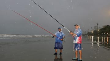 dois homens estão com capa de chuva, em dia chuvoso, pescando na beira do mar. Eles seguram as varas. #paratodosverem