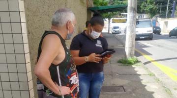 Mulher usando camiseta com identificação da Prefeitura está segurando bloco de notas e conversa com um homem na rua. #Paratodosverem