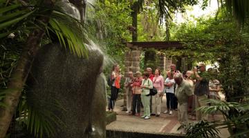 Grupo de turistas está na entrada do orquidário. Em primeiro plano está a ninfa Náiade, de costas. #Paratodosverem