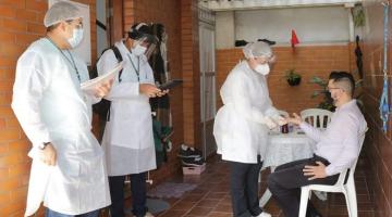 Homem está sentado fazendo teste rápido. Três pesquisadores estão em pé. #Paratodosverem