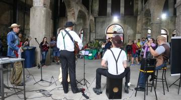 grupo musical está se apresentando em prédio antigo. Músicos estão de costas para a foto e público, de frente. #paratodosverem