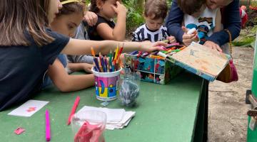crianças se divertem com dobradura e pintura sobre uma mesa. #paratodosverem